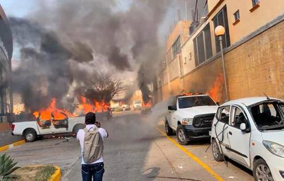 Vehículos en llamas en una calle urbana con una persona observando y humo saliendo del incendio.