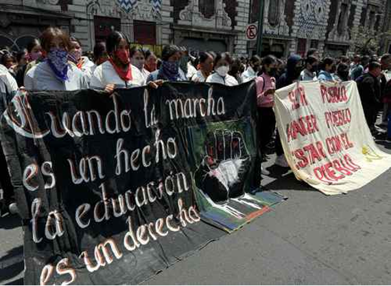 Un grupo de personas con máscaras sostiene dos grandes pancartas con texto en español durante una protesta callejera. Las pancartas hablan sobre los derechos a la educación y la lucha contra la opresión. Se ven edificios al fondo.