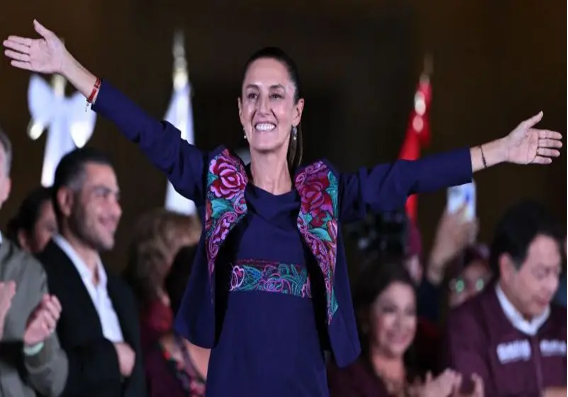 Una mujer está de pie con los brazos extendidos, sonriendo, mientras varias otras personas están al fondo, aplaudiendo y sonriendo.