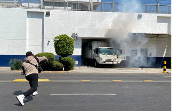 En primer plano, una persona corre, mientras que al fondo, cerca de un edificio, un camión emite humo de su motor.