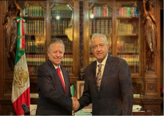 Dos hombres dándose la mano frente a una bandera mexicana.