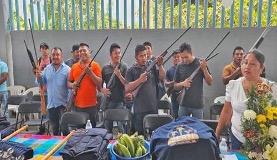 Grupo de hombres sosteniendo rifles mientras estaban parados detrás de una mesa con varios artículos, incluyendo ropa y plátanos, durante una reunión en el interior.