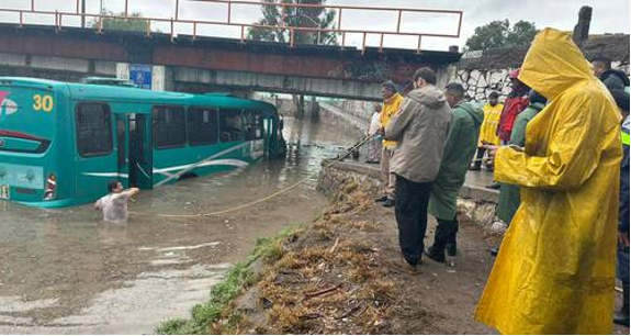 Una calle inundada con un autobÃºs sumergido, gente con impermeables observando y una persona en el agua sujetando una cuerda atada al autobÃºs.