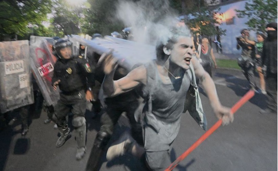 Un manifestante cubierto de polvo blanco corre sosteniendo un poste rojo, perseguido por varios policías con equipo antidisturbios y escudos durante una protesta callejera.