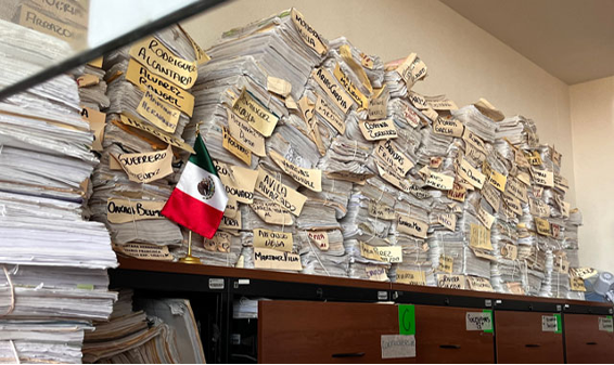 Pilas de documentos en papel etiquetados con diversos nombres se apilan en estantes, con una pequeÃ±a bandera mexicana colocada entre las pilas.