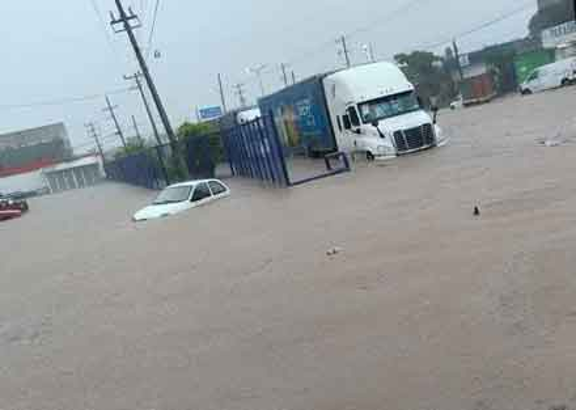 Una calle inundada con vehículos parcialmente sumergidos, entre ellos un automóvil blanco y un camión de grandes dimensiones, en medio de una fuerte lluvia.