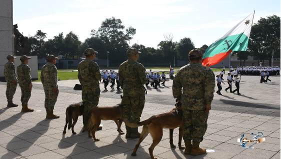 Soldados y perros militares se encuentran en un área pavimentada mientras un grupo de personal uniformado marcha con una gran bandera. Al fondo se ven árboles y otras personas.