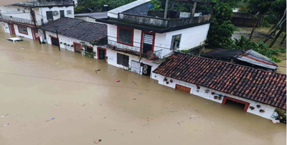 Calle inundada con varias casas parcialmente sumergidas, agua marrón cubriendo la carretera y árboles al fondo.