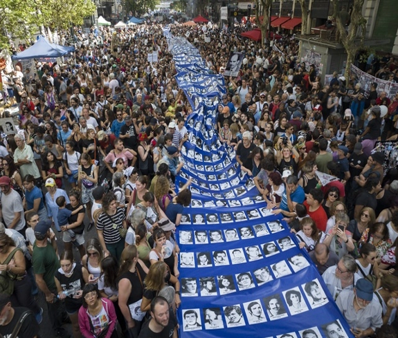 Una gran multitud se reÃºne alrededor de una larga pancarta de tela que muestra rostros, durante un evento pÃºblico al aire libre.