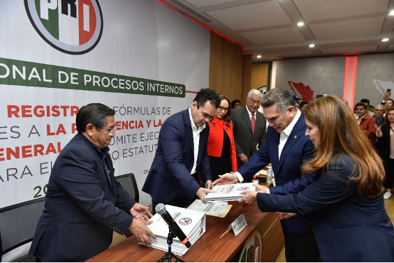 Personas en un ambiente formal intercambian documentos con una pancarta al fondo que muestra las siglas "PRI" y el texto "PROCESOS INTERNOS".