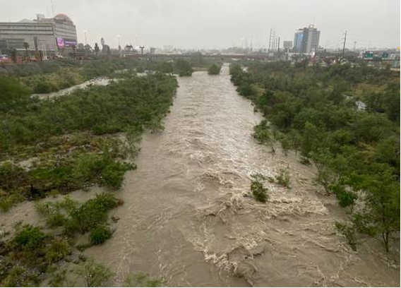 Un rÃo crecido fluye a través de una zona de la ciudad con edificios visibles a ambos lados. El agua está alta y turbulenta, lo que indica fuertes lluvias o inundaciones. La vegetación bordea las riberas del rÃo.