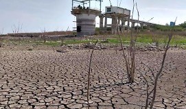 Cauce seco con barro agrietado, con escasa vegetación y una estructura de hormigón con transportadores al fondo.