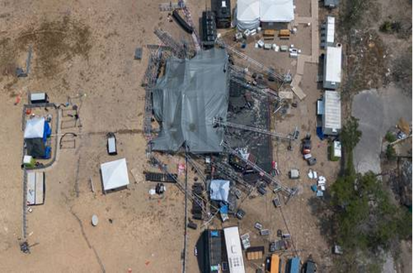 Vista aérea de un evento al aire libre en construcción, con grandes carpas, andamios, contenedores de envío y diversos equipos repartidos en un área abierta de arena. .