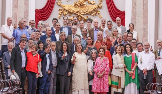 Un gran grupo de personas posando para una fotografía en un ambiente formal con adornos rojos y dorados de fondo. El grupo está formado por hombres y mujeres de distintas edades, vestidos con trajes tradicionales y de negocios.