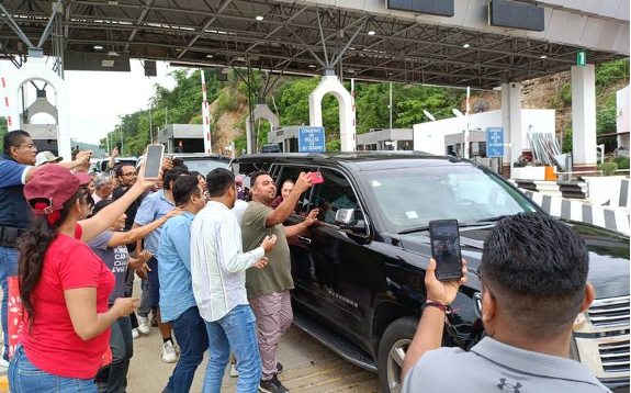 Un grupo de personas se reúne alrededor de una camioneta negra en una cabina de peaje, tomando fotografías y videos con sus teléfonos.