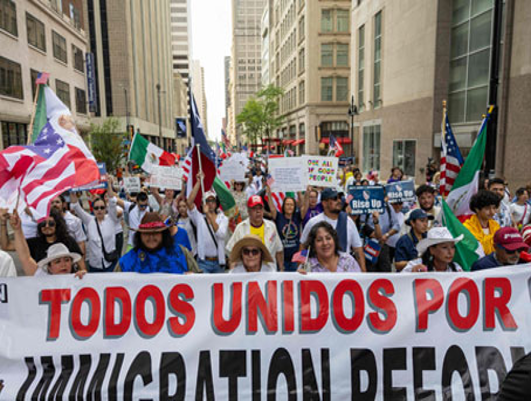 Un grupo diverso de personas participa en una marcha por la reforma migratoria, portando pancartas y banderas. La pancarta dice "Todos Unidos por la Reforma Migratoria" en una calle de la ciudad.