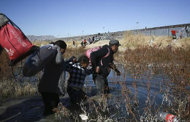 Tres personas caminan por un río poco profundo, cada una con sus mochilas, con pasto y una cerca al fondo. Se ven otras personas a lo lejos.