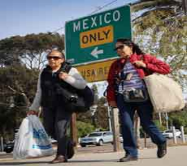 Dos personas pasan junto a un cartel que indica la direcciÃ³n a MÃ©xico, cargando bolsas. Una de ellas lleva gafas de sol y un chaleco negro, mientras que la otra lleva una chaqueta roja. Se ven Ã¡rboles al fondo.