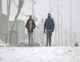 Dos personas caminan por una calle cubierta de nieve, abrigadas con ropa de invierno. Al fondo se ve un coche.