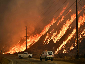 Los bomberos responden a un gran incendio forestal en una ladera junto a una carretera, con dos vehículos de emergencia presentes en primer plano.