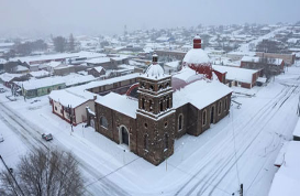 Vista aérea de una ciudad cubierta de nieve con una destacada iglesia de piedra con dos cúpulas. Las calles y los edificios circundantes están cubiertos de nieve bajo un cielo nublado.