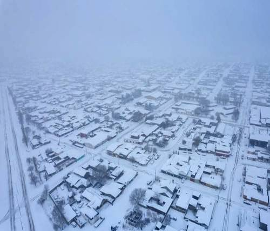 Vista aérea de una zona residencial cubierta de nieve, con casas, calles y una línea de ferrocarril visibles bajo un pesado manto de nieve en un día de niebla.