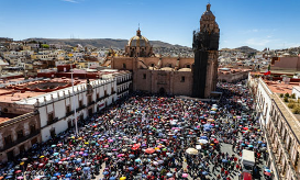 Una gran multitud se reúne en una plaza frente a una iglesia histórica bajo un cielo azul claro, con edificios de la ciudad circundantes y colinas distantes.