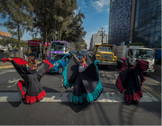 Tres personas con coloridos vestidos tradicionales actúan en un cruce de peatones mientras el tráfico espera, con un telón de fondo de edificios altos y árboles.