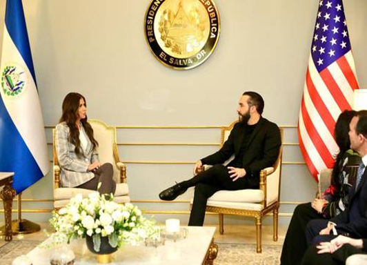Un hombre y una mujer se sientan uno frente al otro en una oficina con banderas de El Salvador y Estados Unidos, debajo de un sello presidencial salvadoreño.
