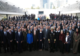 Un gran grupo de personas, incluidos individuos con uniformes navales, posan para una foto al aire libre frente a un edificio moderno.