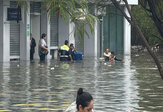 Gente caminando a través del agua inundada hasta las rodillas frente a un edificio con las contraventanas cerradas y un árbol en primer plano.