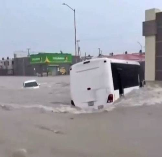 Un autobús y un automóvil blancos parcialmente sumergidos en las aguas de la inundación en una calle, con un edificio y una señalización visibles en el fondo.