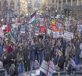 Una multitud grande y diversa sostiene carteles y banderas durante una protesta al aire libre en una calle de la ciudad.