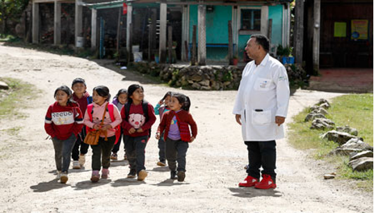 Un hombre con bata blanca se encuentra de pie en un camino de tierra con un grupo de niños pequeños que llevan mochilas. Están al aire libre con un edificio al fondo.