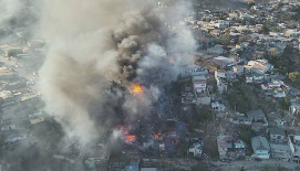 Vista aérea de un gran incendio en una zona urbana, con humo espeso y llamas visibles extendiéndose entre los edificios.