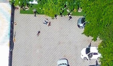 Vista aérea de un área de estacionamiento con algunos autos estacionados, gente caminando y árboles verdes que brindan sombra.