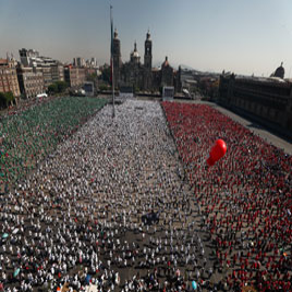 Una gran multitud en una plaza pública forma los colores de la bandera mexicana con su ropa. Un solitario globo rojo flota sobre la multitud. Al fondo se ven edificios y una catedral.