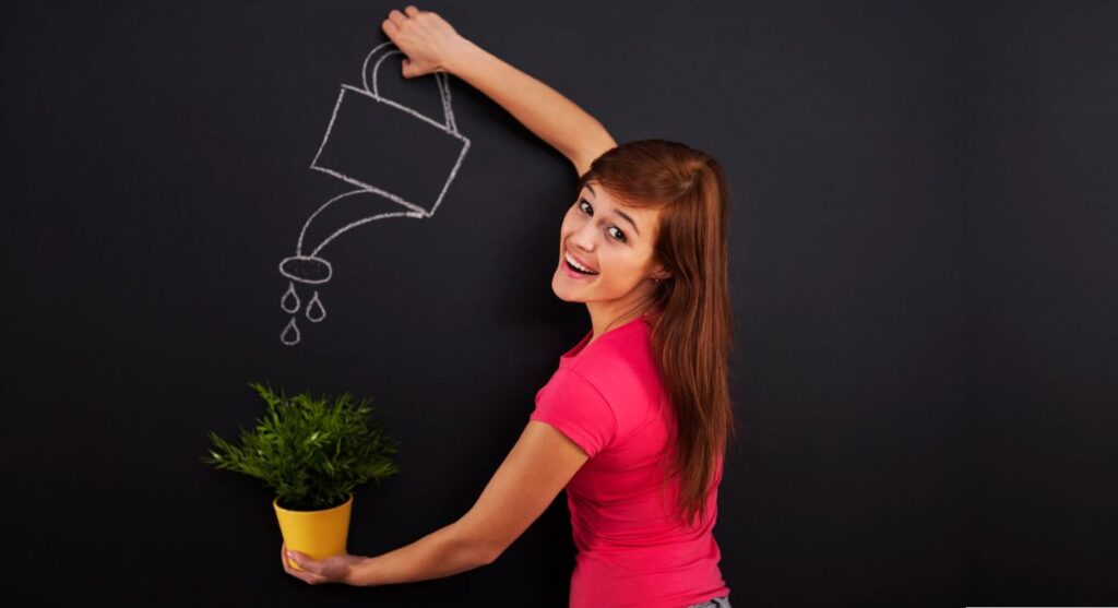 Una mujer con una camisa rosa sostiene una planta en maceta debajo del dibujo de una regadera en una pizarra, sonriendo a la cámara, un sÃmbolo de la resiliencia mientras nutre el crecimiento con cuidado.