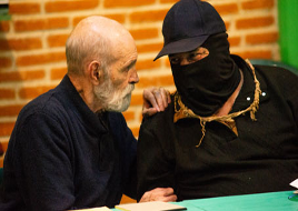 Un hombre mayor, de barba gris, habla de cerca con una persona que lleva una máscara negra, gorra y ropa oscura, sentada en una mesa con una pared de ladrillos al fondo.