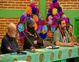 Cuatro personas estÃ¡n sentadas en una mesa decorada con micrÃ³fonos; al fondo hay flores de papel de colores y una pared de ladrillos.