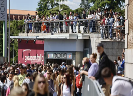 Una gran multitud de personas se encuentra de pie y caminando afuera de un edificio, algunos en una pasarela superior y otros en el nivel del suelo.