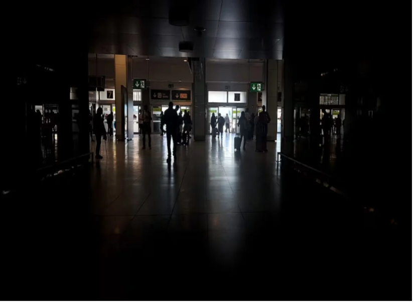 Siluetas de personas caminando por un espacio interior poco iluminado con seÃ±ales de salida iluminadas y ventanas en el fondo.