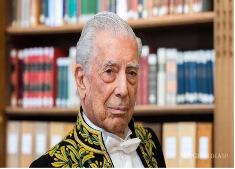 Un anciano con un traje formal con bordados dorados se encuentra frente a una estantería llena de libros.