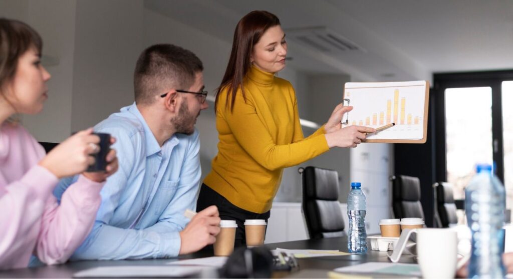 Una mujer con un suéter amarillo presenta un gráfico de barras en un portapapeles a dos colegas en una mesa de conferencias, mientras discuten sobre las startups mientras beben y comparten papeles.