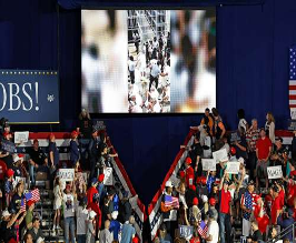 Una multitud en un mitin polÃ­tico en un lugar cerrado permanece frente a una pantalla grande que muestra una imagen borrosa; algunas personas sostienen carteles y visten ropa de color rojo, blanco y azul.