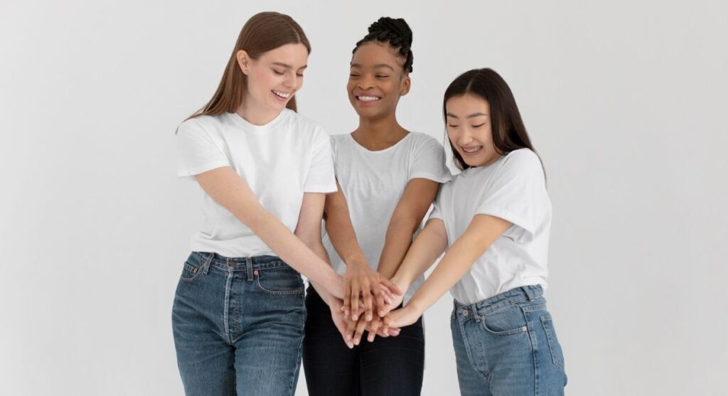 Tres mujeres, juntas, sonriendo y uniendo las manos en el centro, representan la hermandad y la sororidad. Llevan camisetas blancas y vaqueros sobre un fondo blanco liso.