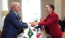 Dos personas se dan la mano sobre una mesa con pequeñas banderas brasileñas y mexicanas, mientras otros se sientan y observan en una habitación con cortinas.