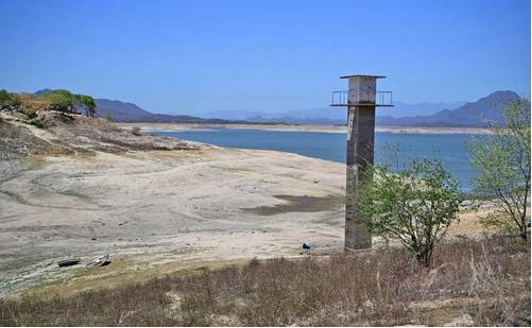 Una torre alta y estrecha se alza sobre una tierra seca y árida cerca de un gran cuerpo de agua bajo un cielo azul claro, con montañas distantes en el fondo.