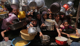 Una multitud de personas sostiene ollas y cuencos para alcanzar la comida que se sirve al aire libre, lo que sugiere un evento de ayuda humanitaria o de distribución de alimentos.