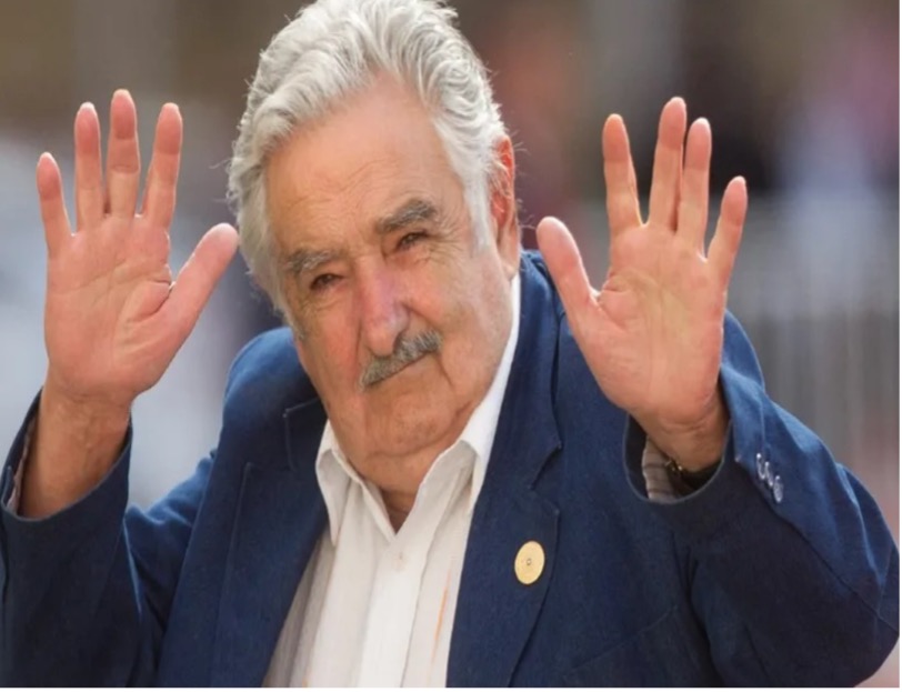 Un hombre mayor, de cabello gris y bigote, viste un blazer azul y una camisa blanca, levanta ambas manos con las palmas abiertas y mira a la cÃ¡mara.